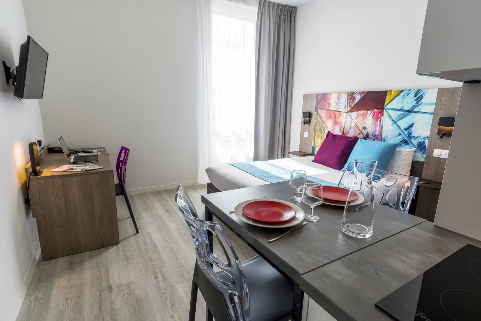 Modern hotel room apartment with double bed, dining table set with red plates and glasses, desk with purple chair, colorful headboard and large window. Bordeaux.