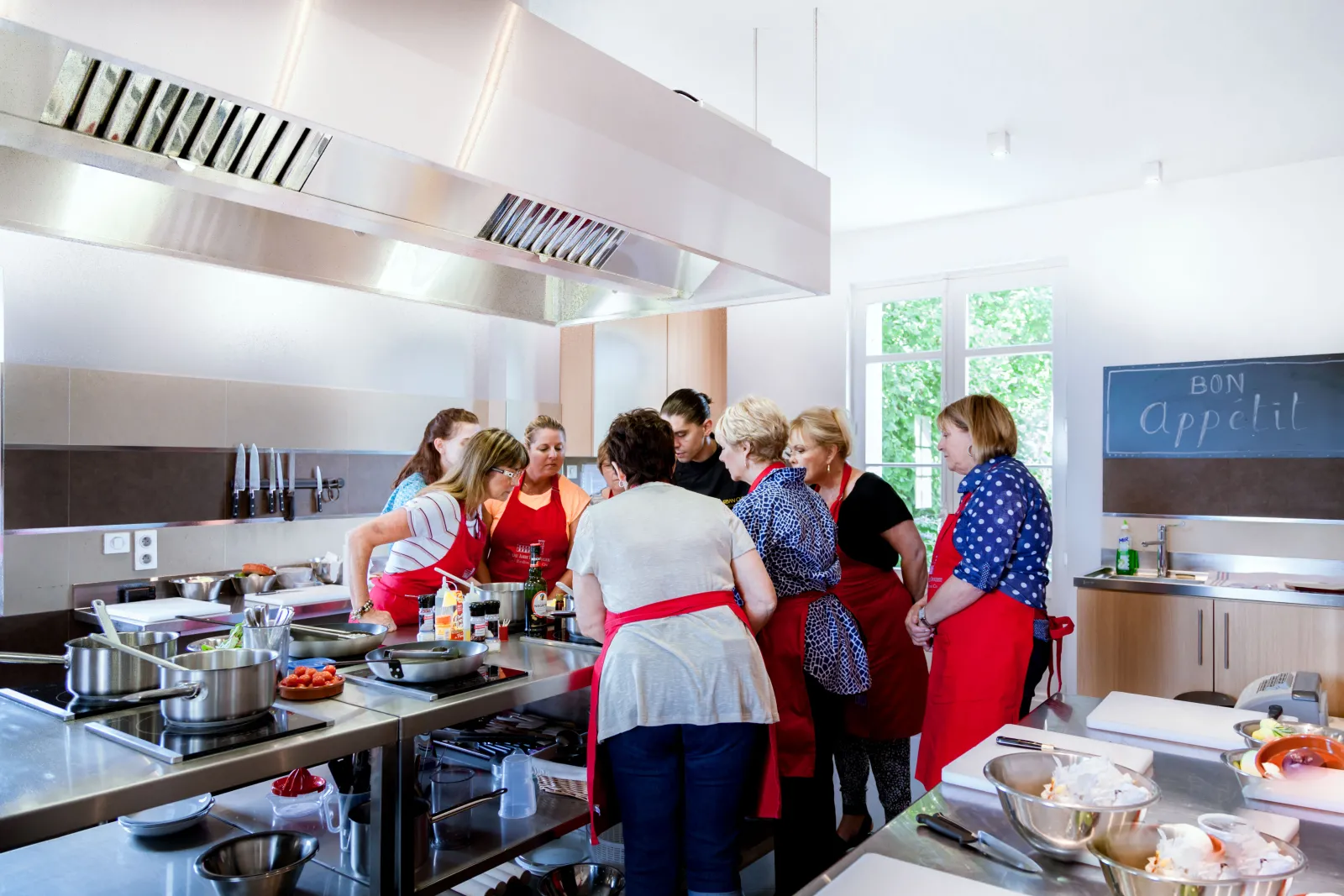 Group of women wearing red aprons taking a cooking class in a modern professional kitchen at Château Ambe Tour Pourret in Bordeaux.