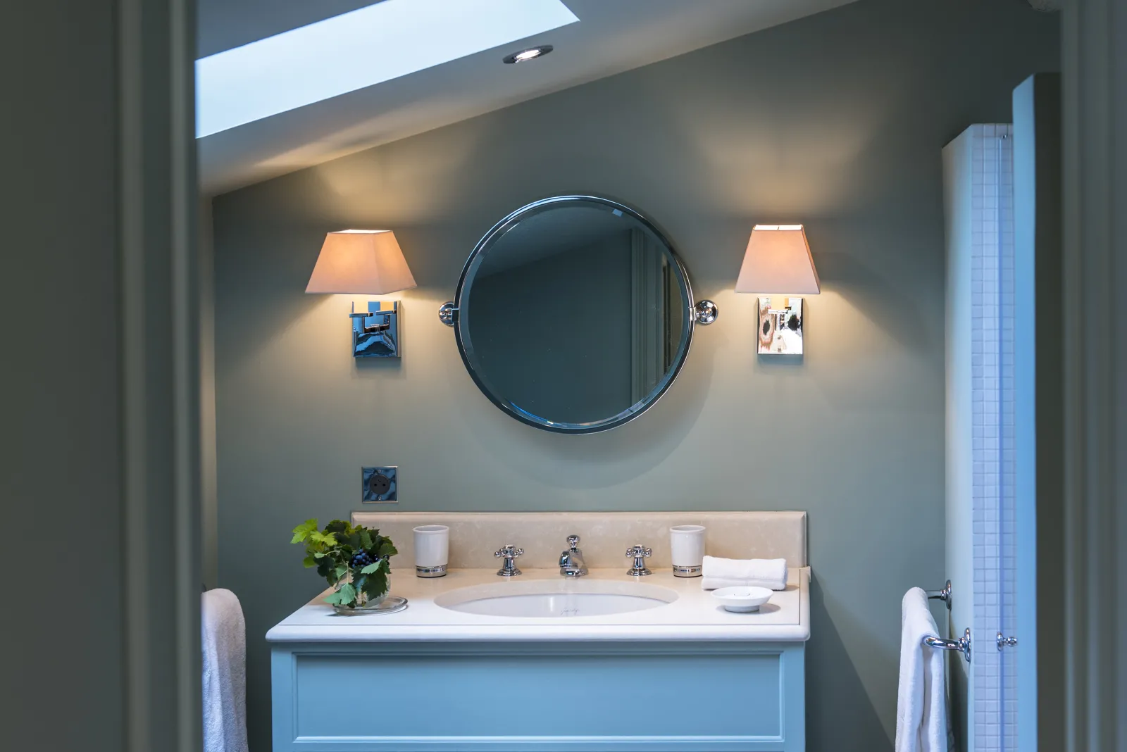 Modern bathroom featuring a white sink, round blue mirror, two wall sconces with beige shades, skylight ceiling, and minimalist décor.