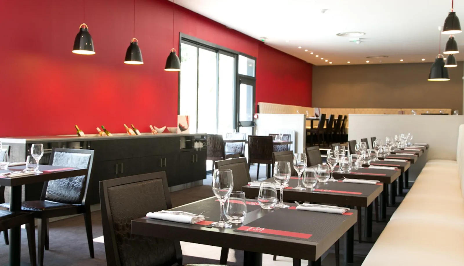 Modern restaurant dining room at Mercure Bordeaux-Lac hotel featuring red and beige walls, black tables set with glassware and cutlery, black chairs, pendant lights and large windows