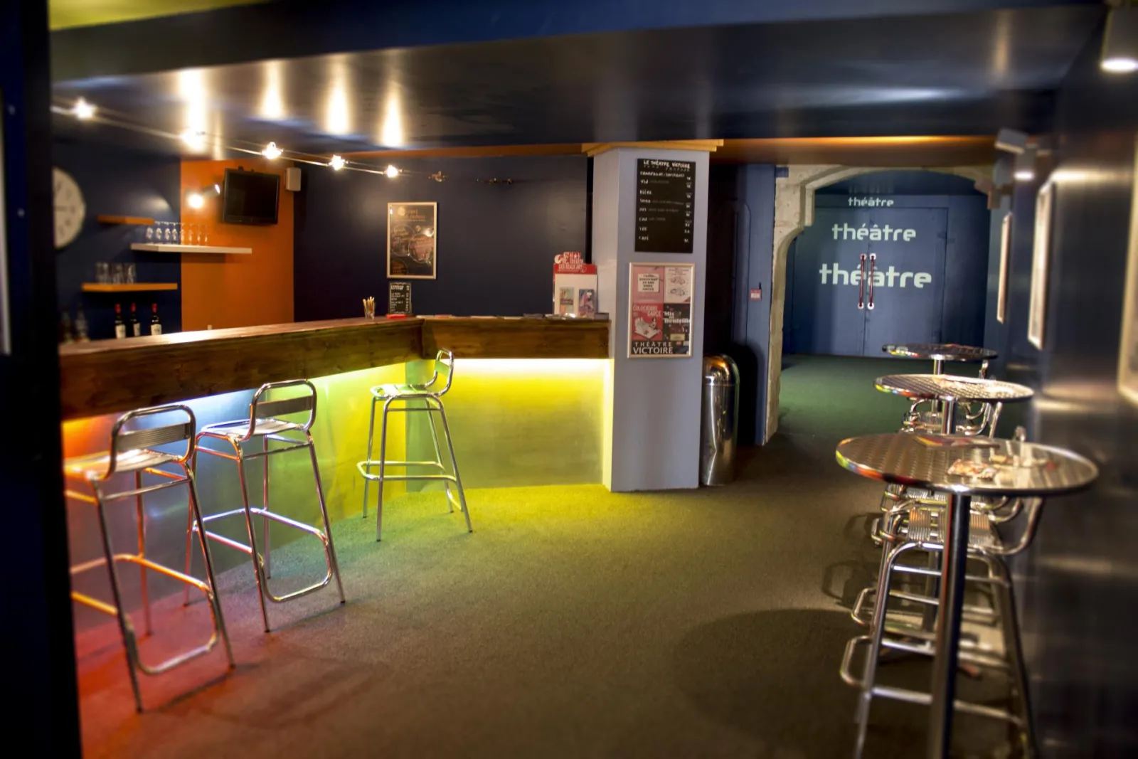 Interior of Théâtre Victoire in Bordeaux featuring a modern bar with yellow neon lighting, high stools, round tables, and the theatre entrance in the background