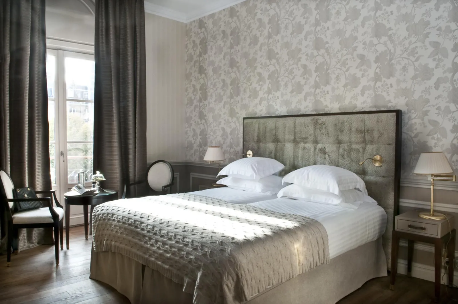 Elegant bedroom at Hôtel de Sèze in Bordeaux featuring a double bed with white pillows, grey upholstered headboard, floral patterned wallpaper, long grey drapes, city-view window, gold bedside lamps and classic furniture