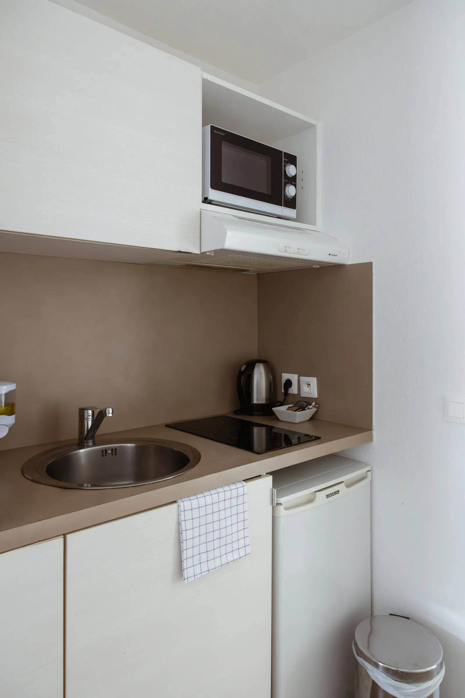 Compact kitchenette with beige countertop, round sink, built-in microwave, white dishwasher, kettle and kitchen accessories in a serviced apartment hotel in Bordeaux-Mérignac