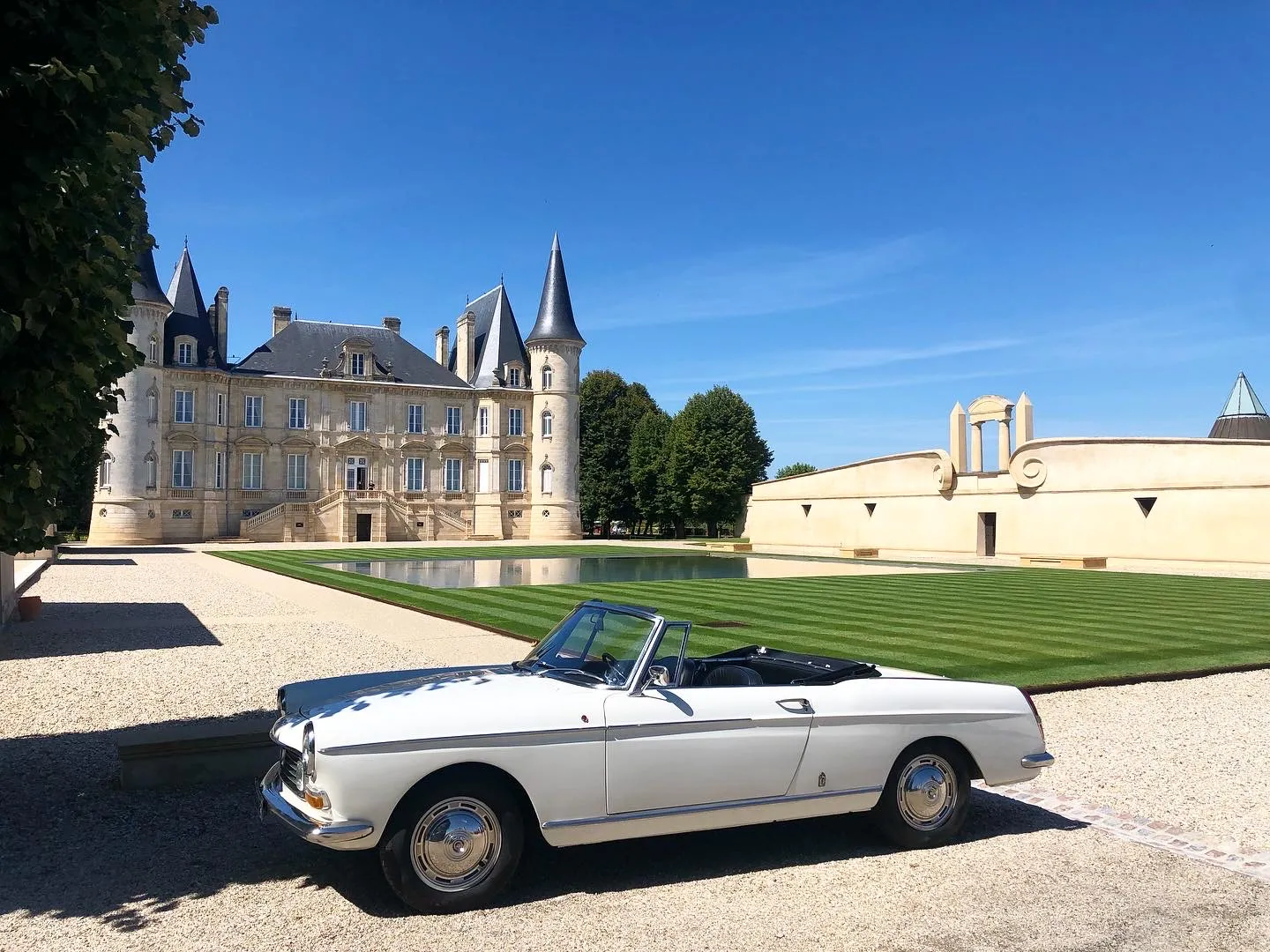 White and blue convertible from the 1950s parked in front of a French château with manicured grounds, striped lawn and reflecting pool.