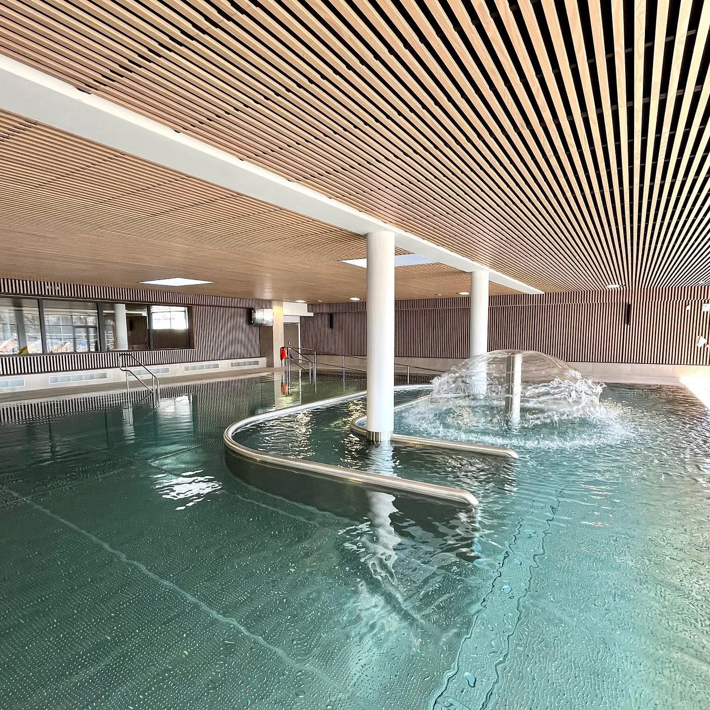 Interior of UCPA Aqua Stadium aquatic center in Mérignac near Bordeaux, featuring two turquoise pools, white columns, wooden slat ceiling, water jets and natural lighting.