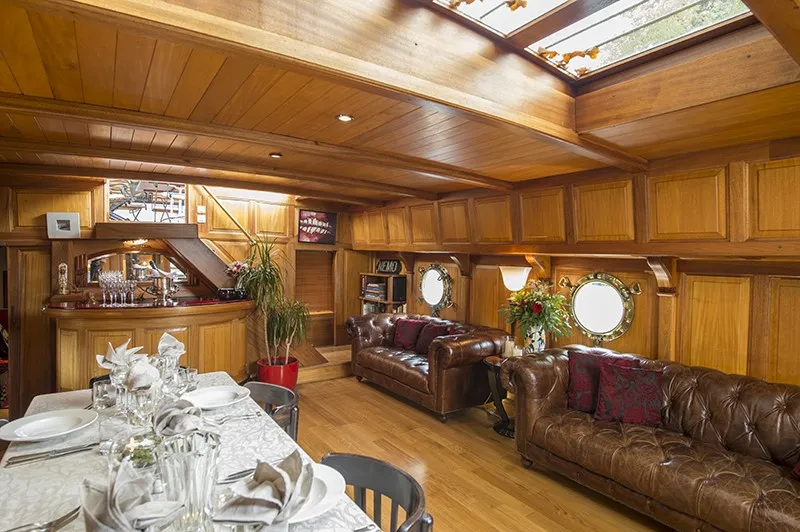 Luxurious wooden yacht interior featuring an elegant lounge with brown leather sofas, formal dining table, rich wood paneling, and round portholes