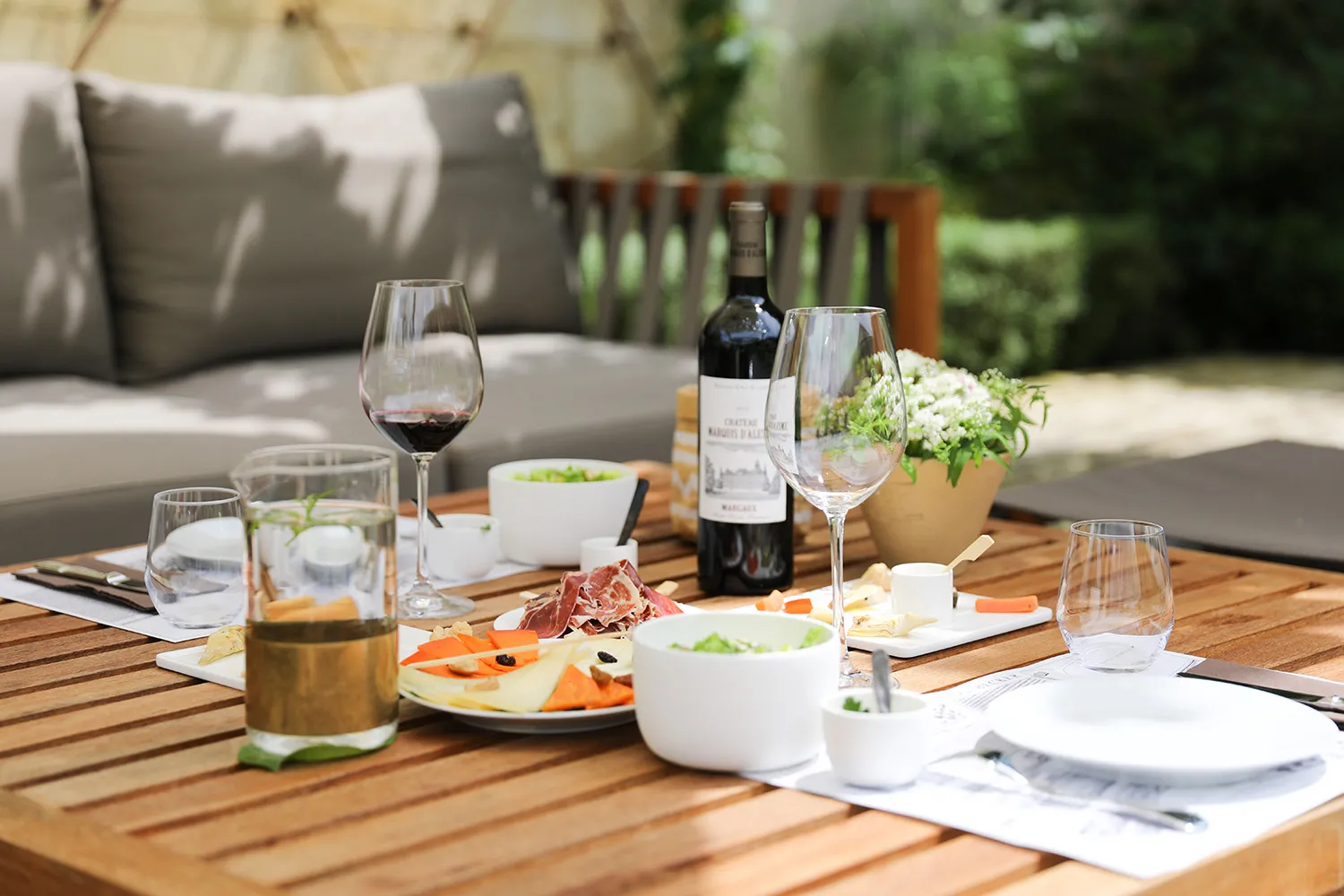 Elegant wooden dining table set with wine glasses, gourmet plates, Bordeaux wine bottle and flowers, outdoor terrace with upholstered seating.