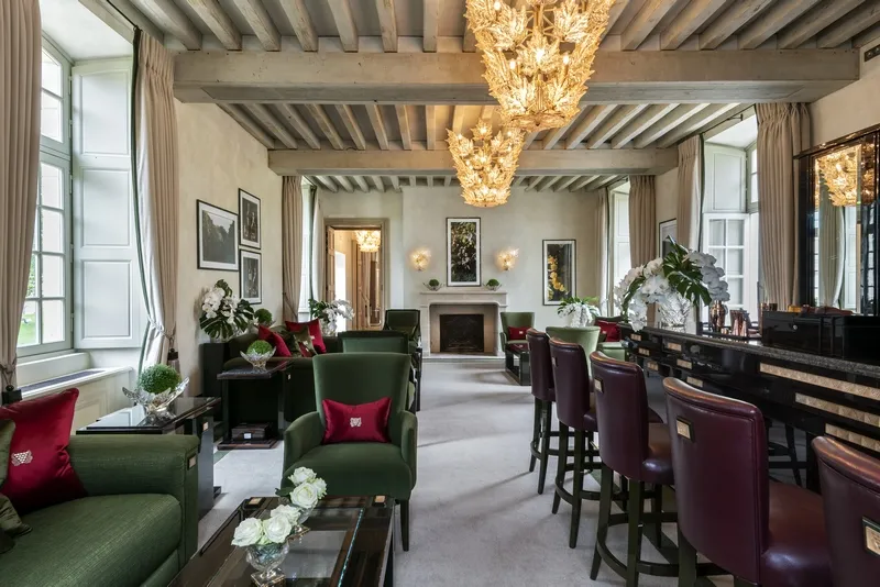 Elegant grand lounge of Hôtel Lalique featuring exposed wooden beams, green and burgundy armchairs, crystal chandelier, fireplace, bar counter, and grand piano in sophisticated classic décor.