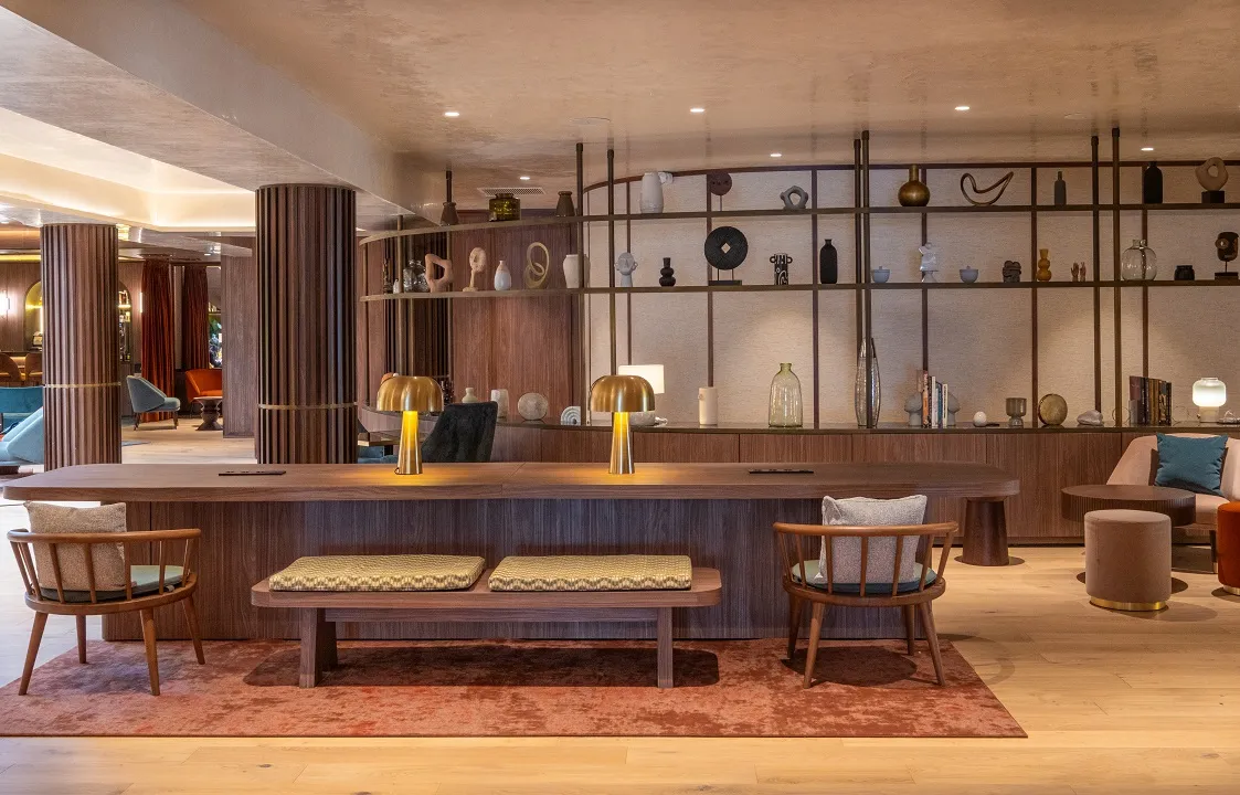 Elegant reception area of Hôtel Burdigala featuring wooden counter, rattan chairs, upholstered bench, brass lamps and minimalist decorative shelving in Bordeaux