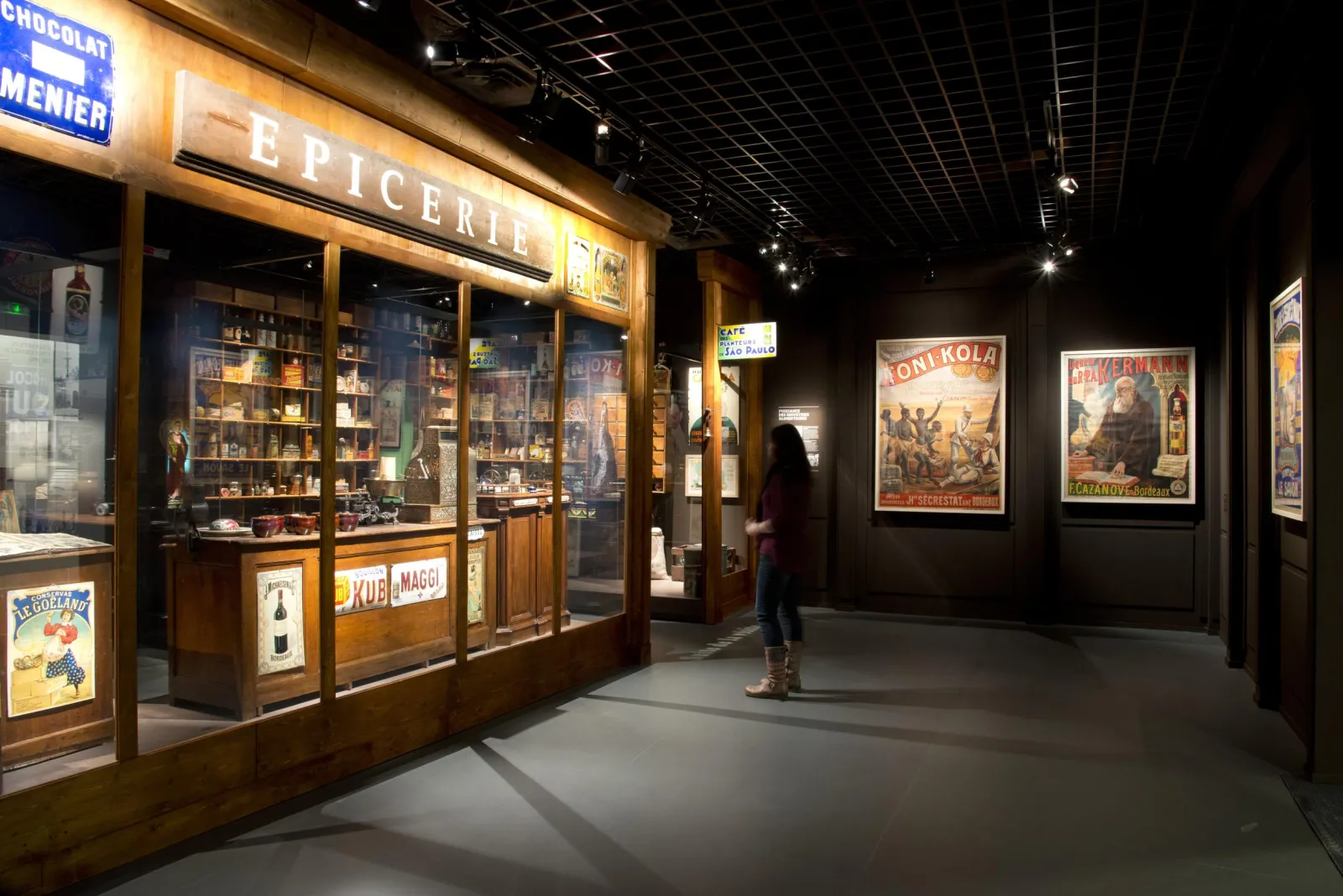 Museum display at the Musée d'Aquitaine in Bordeaux featuring a historical grocery store with vintage products and advertising posters, a visitor viewing the wall exhibitions