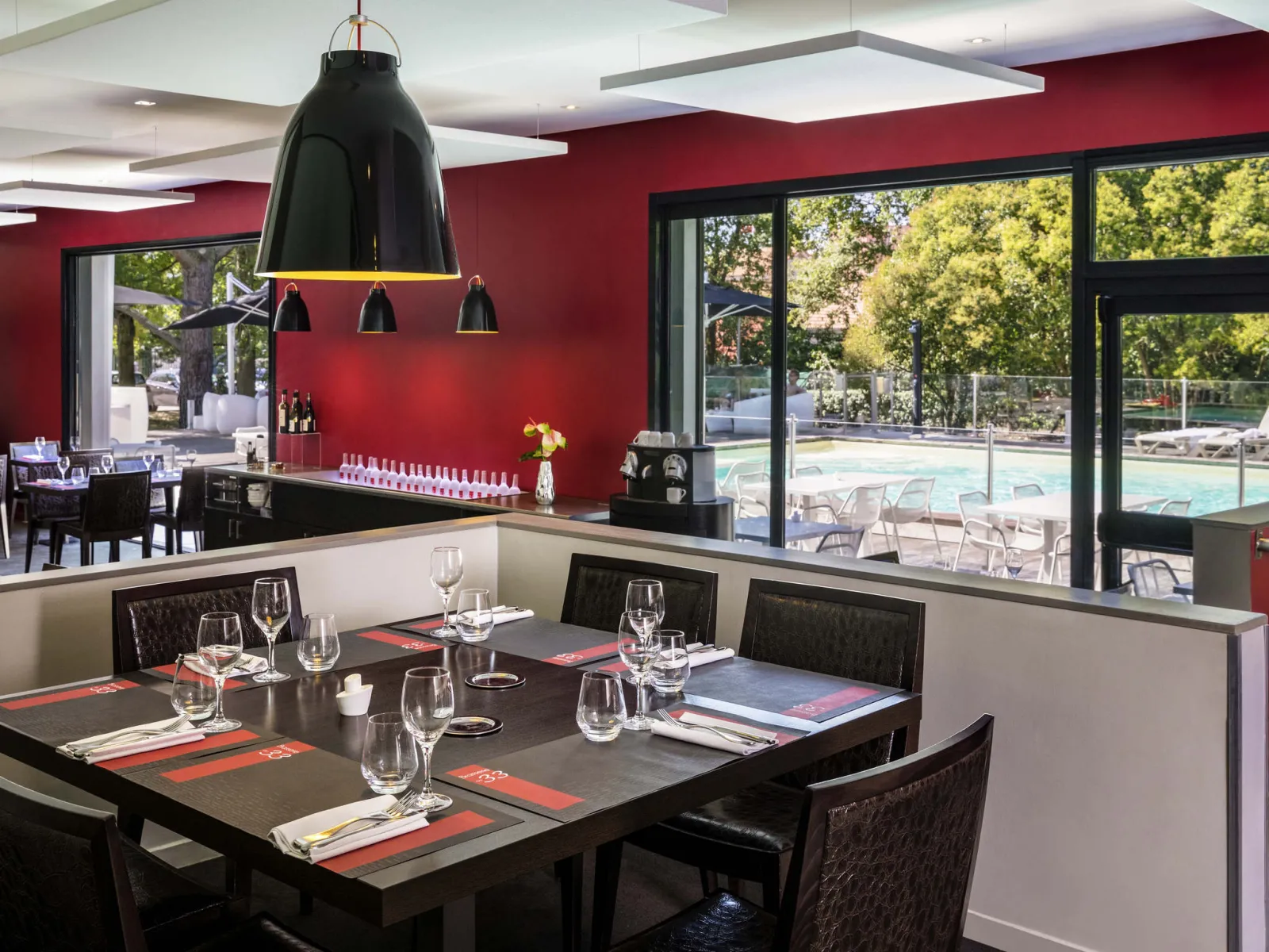 Modern restaurant featuring vibrant red walls, elegantly set black dining tables with glassware and place settings, dark pendant light fixture, and floor-to-ceiling windows overlooking a swimming pool and outdoor lounge area.