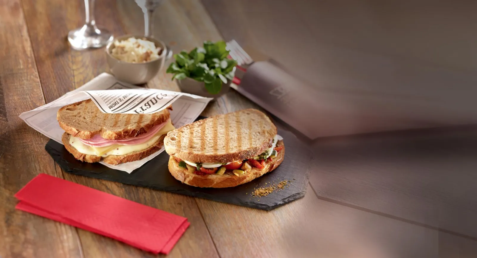 Two gourmet toasted sandwiches on a black slate board at Ibis Kitchen Lounge in Bordeaux, served with salad, red napkin and wine glasses
