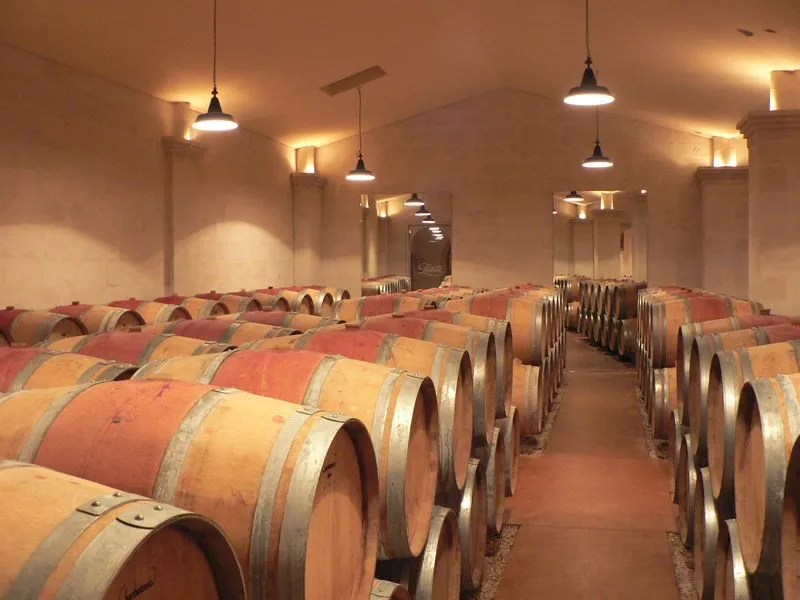 Wine cellar at Château Lestrille in Bordeaux featuring rows of oak barrels lined up under a ceiling lit by pendant lamps.