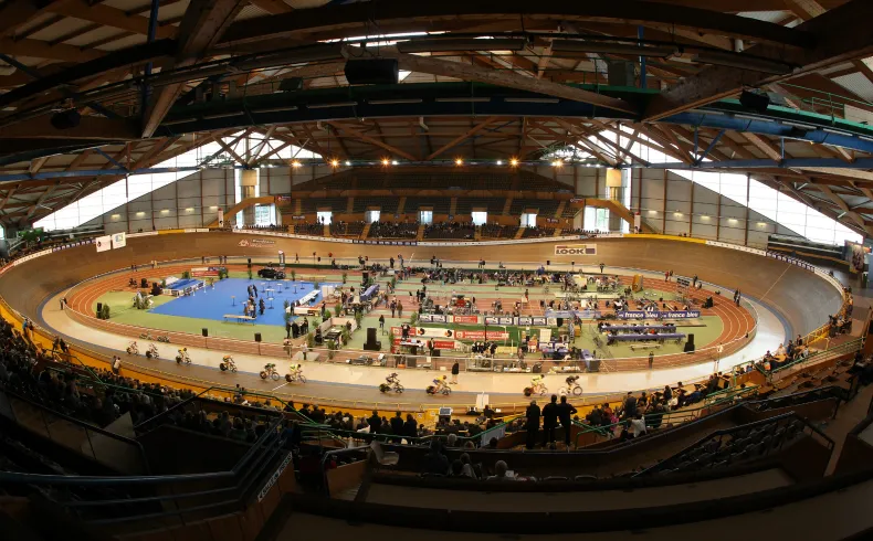 Panoramic view of Bordeaux velodrome featuring its red clay oval cycling track, packed spectator stands, and distinctive wooden and glass architecture with an arched roof.
