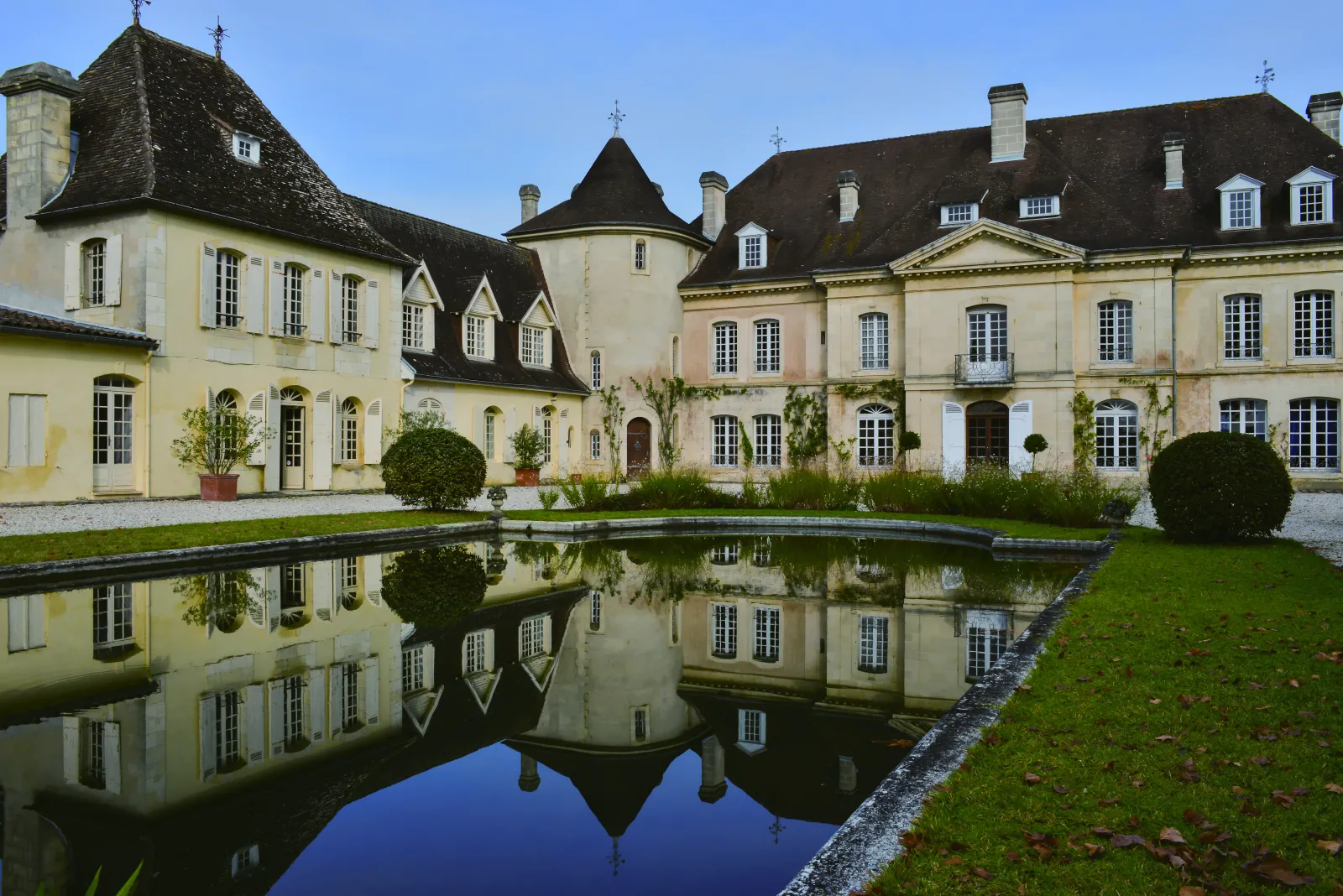 Château Bouscaut in Bordeaux, elegant light stone facade with slate roofs, reflected in a calm water basin, surrounded by landscaped gardens and greenery.