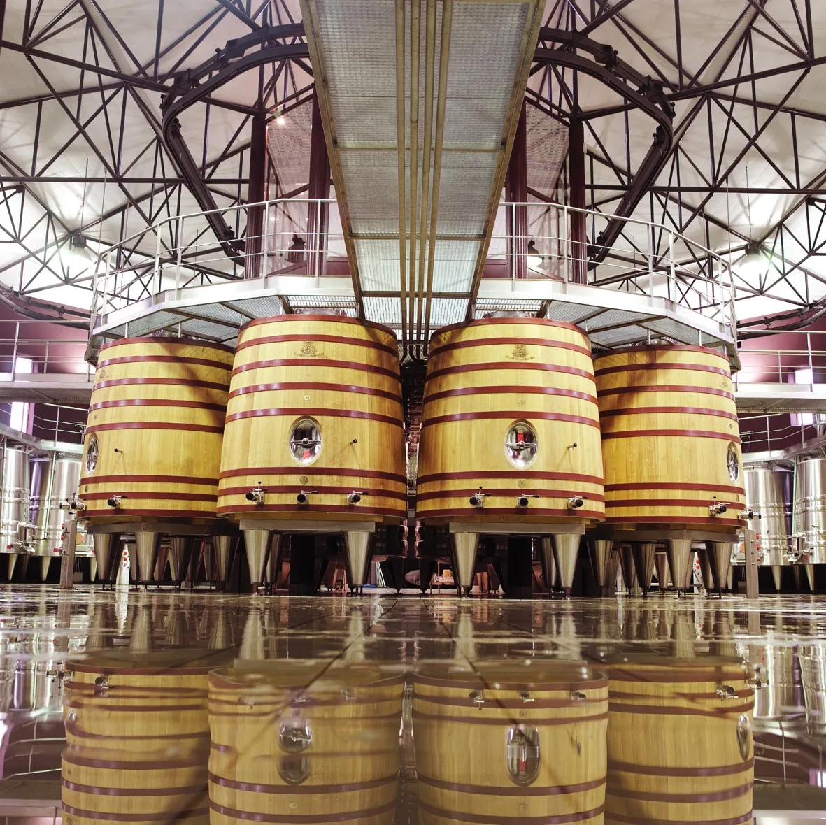 Interior of a winemaking cellar featuring four large wooden vats with red and gold hoops, symmetrically arranged beneath an industrial metal framework. The polished floor mirrors the barrels in perfect reflection.
