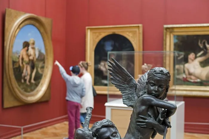 Interior of the Bordeaux Museum of Fine Arts featuring a bronze angel sculpture in the foreground and a visitor photographing paintings displayed on red walls.