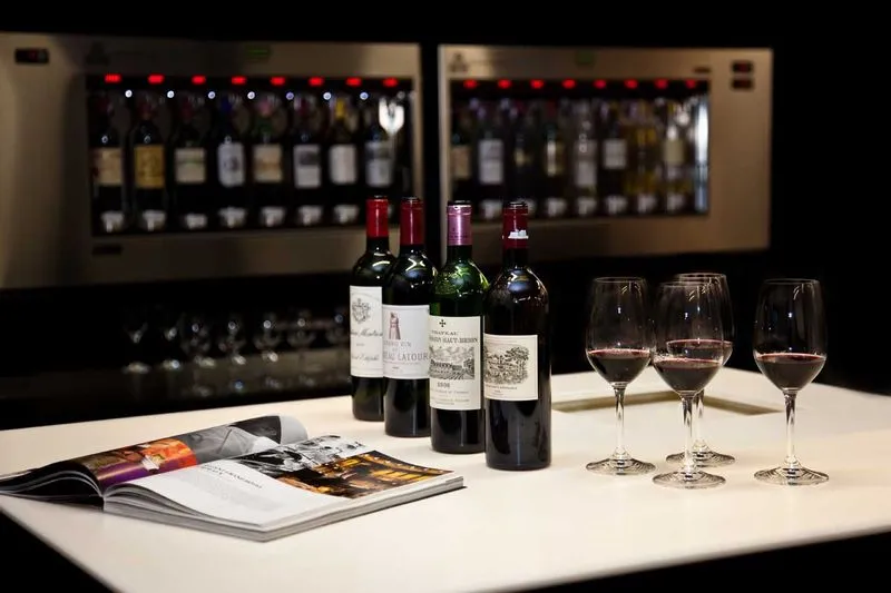 Wine tasting at Max Bordeaux Wine Gallery featuring Bordeaux bottles, filled wine glasses, and wine documentation in front of illuminated wine storage racks