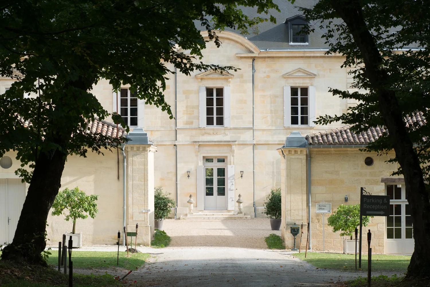 Elegant facade of a classic beige stone building in Bordeaux, featuring columns framing the main entrance, symmetrical windows, and a mansard roof, surrounded by green trees.
