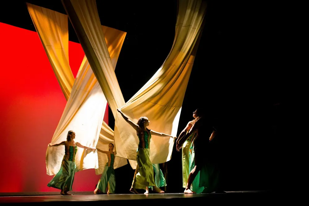 Five dancers in green dresses manipulate large golden and white fabrics on stage with a red background, creating a dynamic artistic performance at the Haillan Warehouse in Bordeaux.
