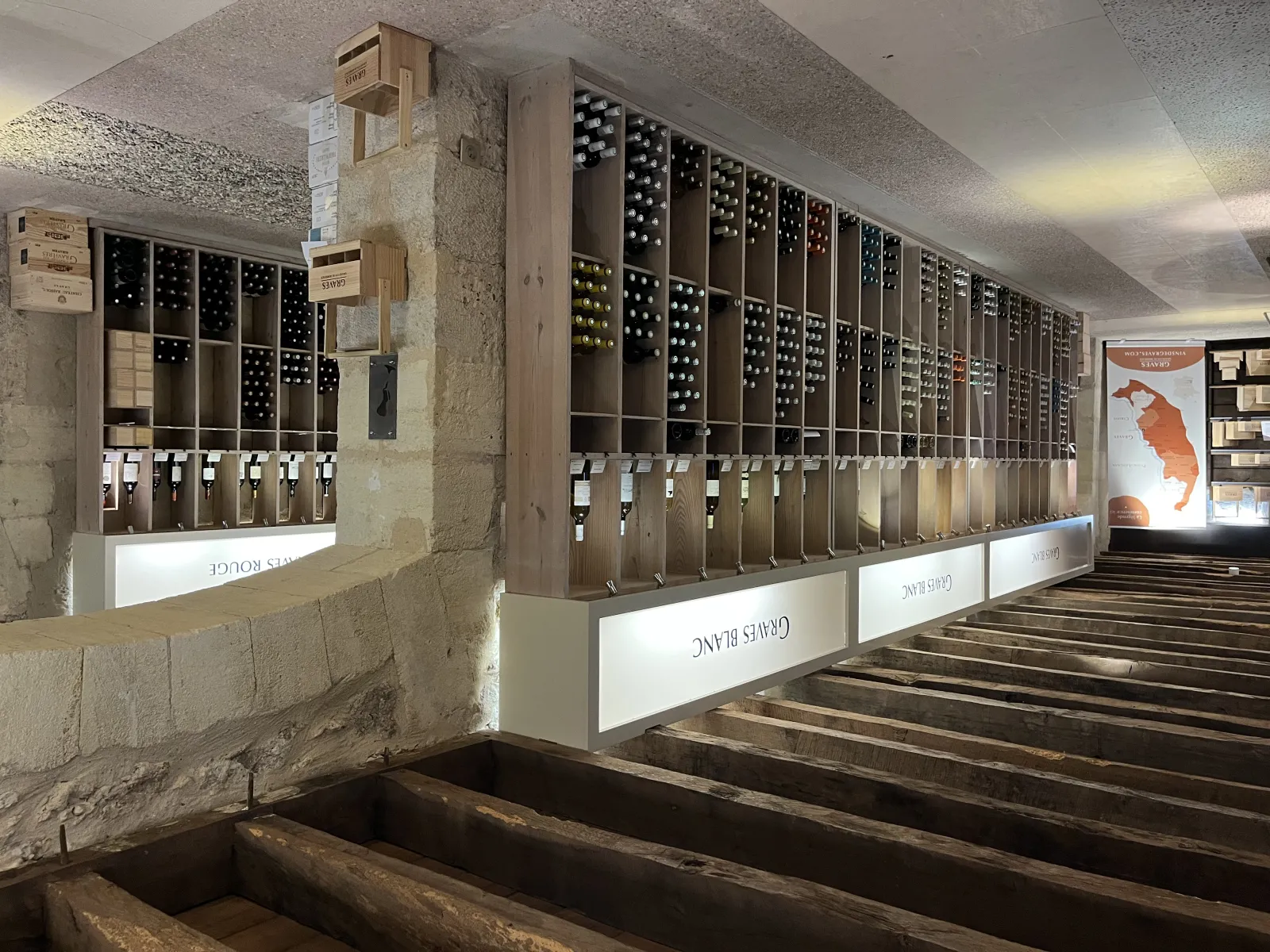 Underground wine cellar at the Maison des Vins de Graves in Bordeaux featuring wooden wine racks with bottles, exposed stone walls, and modern lighting.