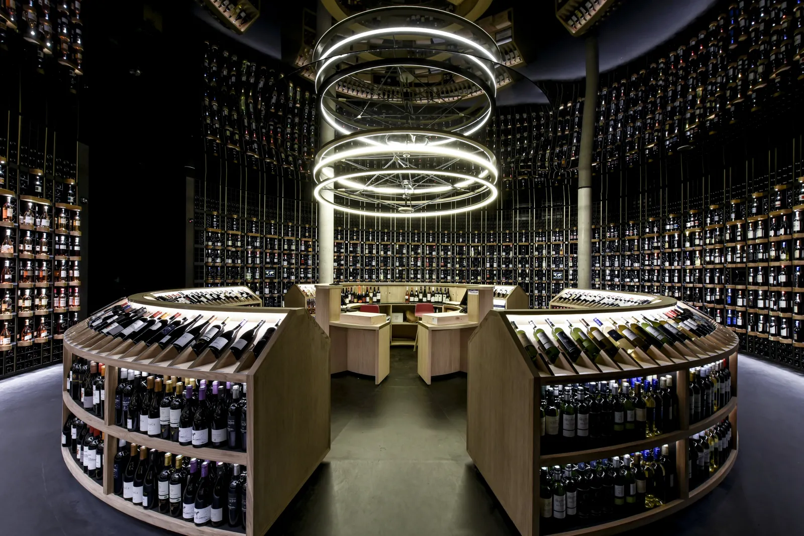 Modern wine cellar of Latitude 20 Wine Bar in Bordeaux, featuring curved shelves filled with wine bottles, a central wooden counter, and circular white ceiling lighting.