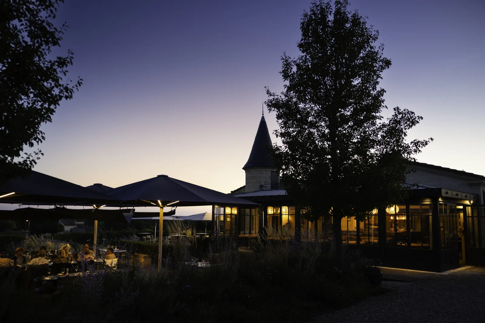 Night view of Château de Candale in Bordeaux featuring its distinctive lit tower, glass buildings, and covered terrace with dining guests under canvas awnings at twilight.