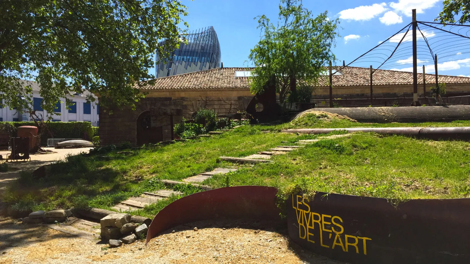 Les Vivres de l'Art, a cultural venue in Bordeaux featuring historic buildings, modern metal structures, green lawn and trees, with the Cité du Vin visible in the background.
