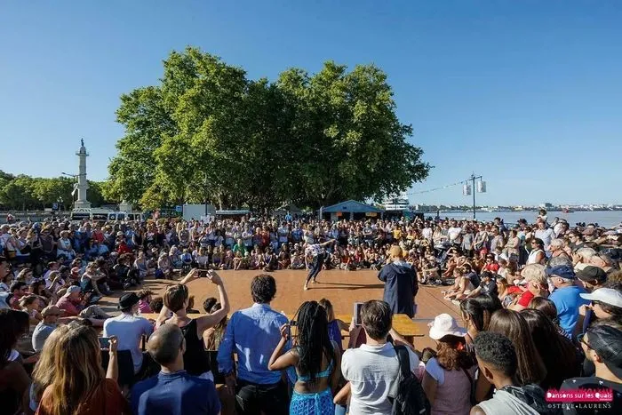 Large crowd of people gathered on the Bordeaux waterfront for an outdoor dance festival event, with trees, a monument, and the port visible in the background under a clear blue sky.