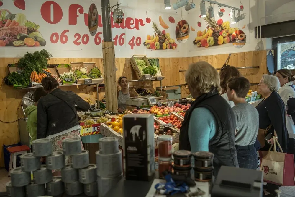 Interior of the Chartrons covered market in Bordeaux with customers shopping at a fresh produce stall, wall display with fruits and phone number