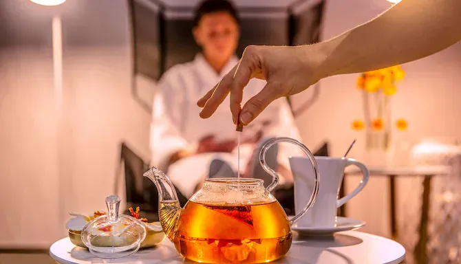 A hand pouring hot tea from a glass teapot into a cup at a spa in Bordeaux, with a professional in the background