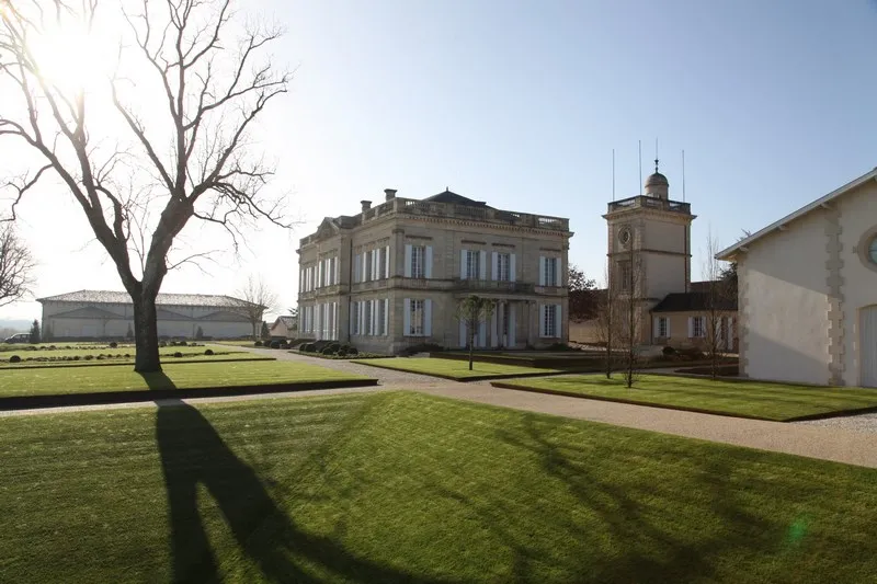 Overview of Château Gruaud Larose in Bordeaux featuring the main stone façade, a clock tower, a bare tree in the foreground, and outbuildings set within a green park.