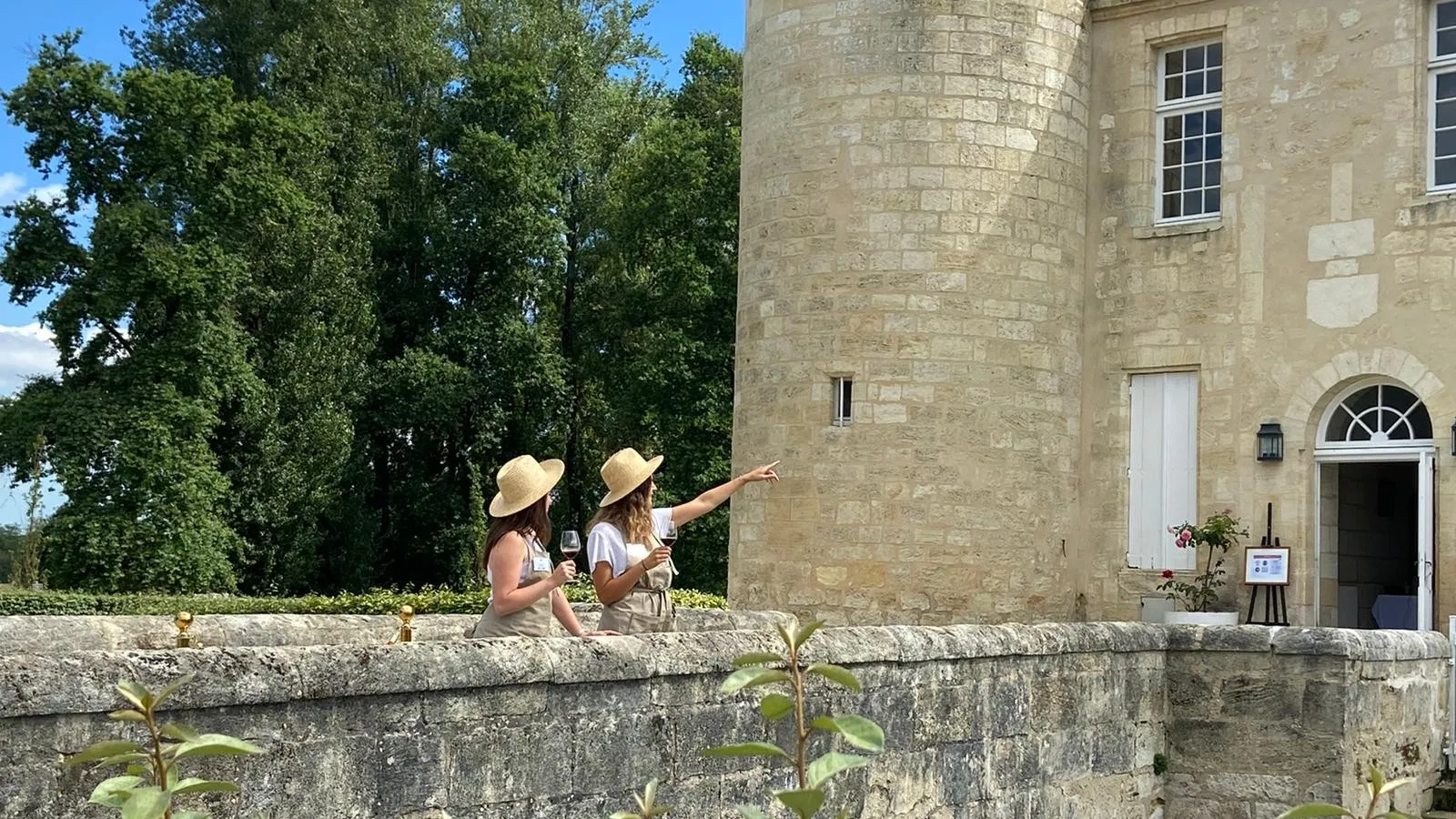 Two women wearing straw hats enjoy wine tasting on the stone terrace of Château d'Agassac in Bordeaux, with the stone facade and lush gardens in the background.