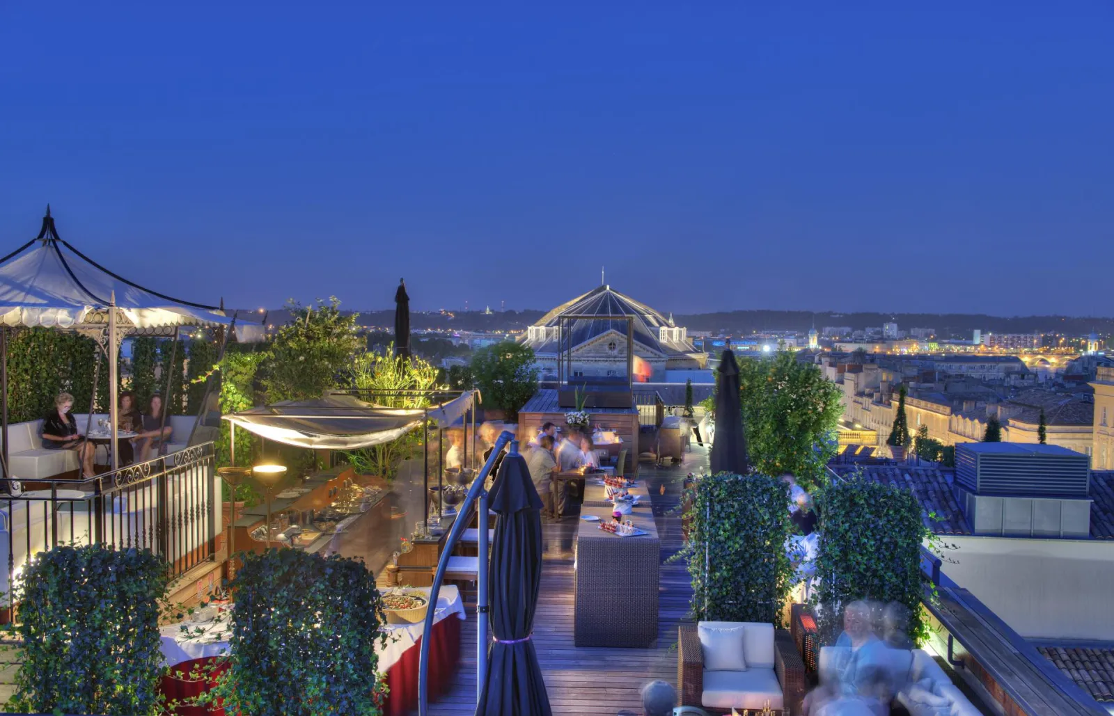 Luxury rooftop terrace of Intercontinental Grand Hotel Bordeaux at dusk, featuring panoramic city views, designer furniture, landscaping, and iconic domed architecture in the background.