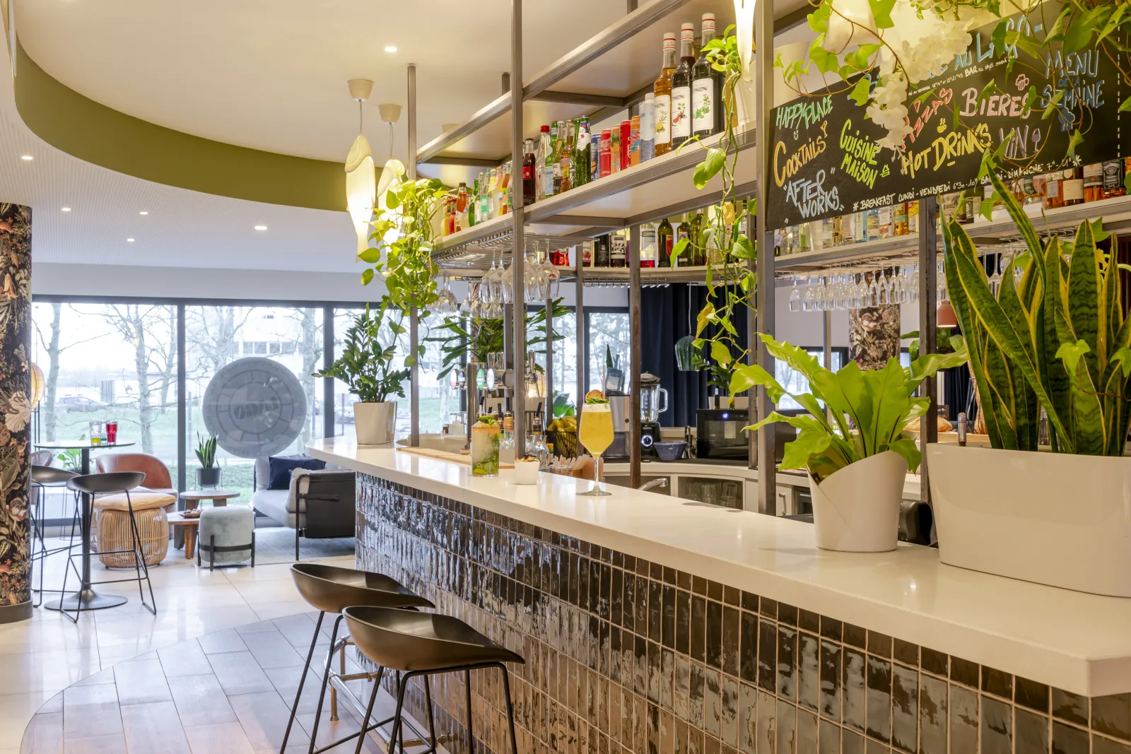 Modern interior of Lago bar in Bordeaux featuring a black and white tiled counter, bottle shelves, green plants, high stools, and large windows overlooking the city.