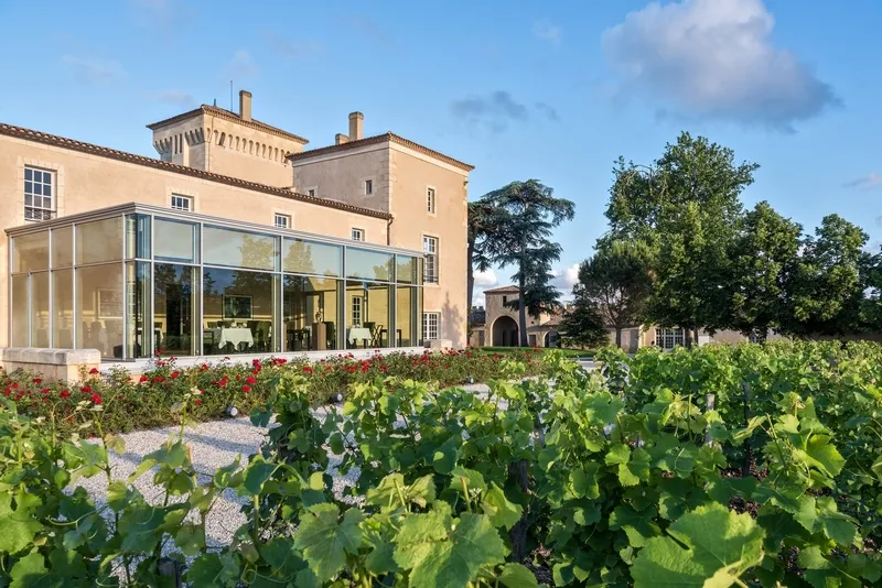 Exterior view of the Lalique restaurant at Château Lafaurie Peyraguey in Bordeaux, featuring a modern glass extension overlooking vineyards, with historic stone buildings and blue sky
