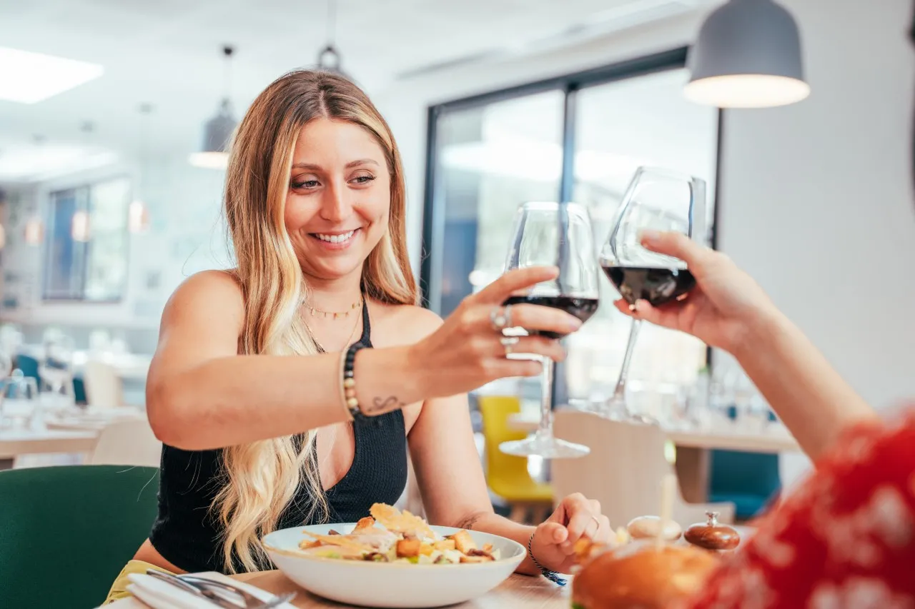 Two women clinking wine glasses during a meal at a modern restaurant in Holiday Inn Bordeaux Mérignac, with a salad and fresh fruits on the table.