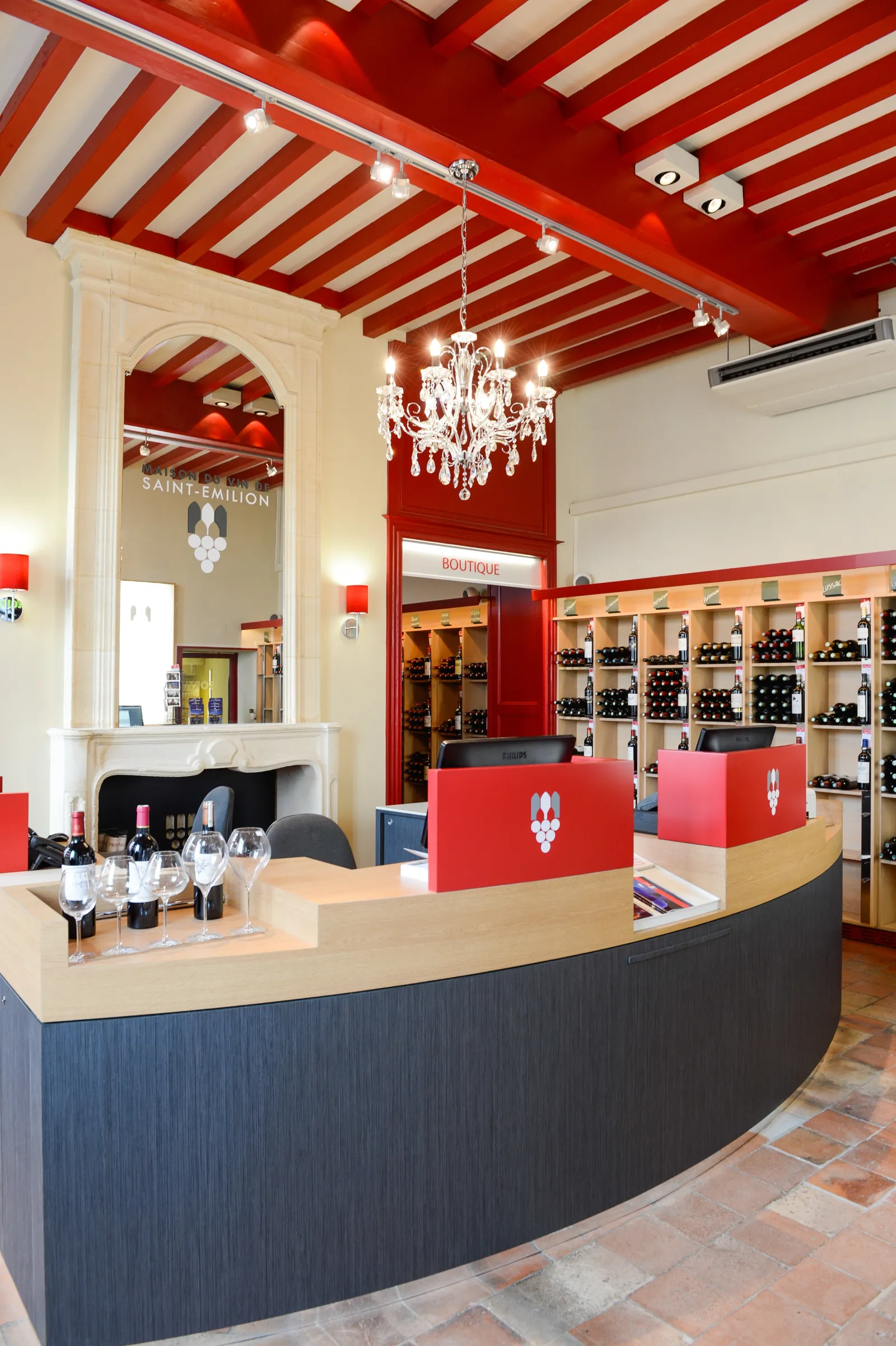 Elegant interior of the Maison du Vin de Saint-Emilion in Bordeaux featuring a curved tasting counter, red and white beamed ceiling, crystal chandelier, wine bottle shelves, and a decorated fireplace.