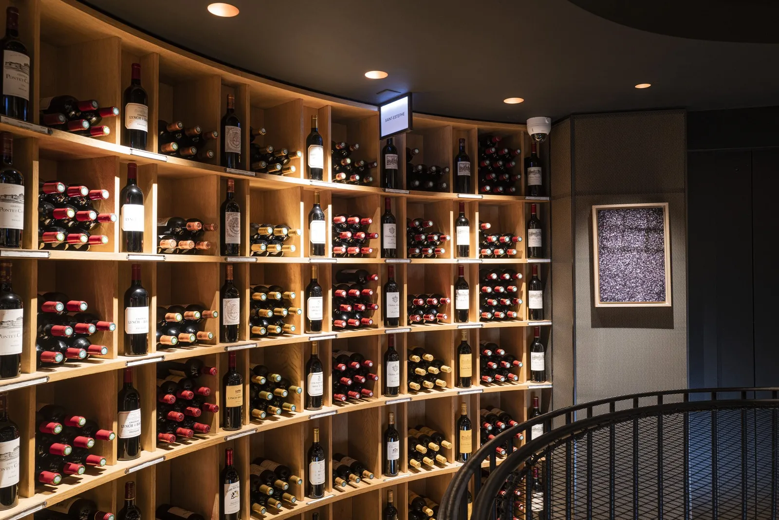 Modern wine cellar with illuminated light wood shelving displaying red wine bottles and colored capsules, interior of L'Intendant restaurant in Bordeaux.