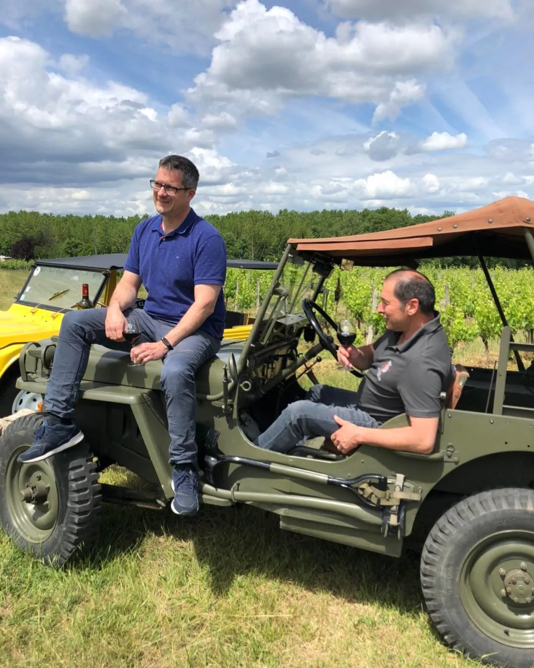Two men chatting on vintage military Jeeps in the vineyards of Château Lagrange near Bordeaux, with cloudy sky overhead.