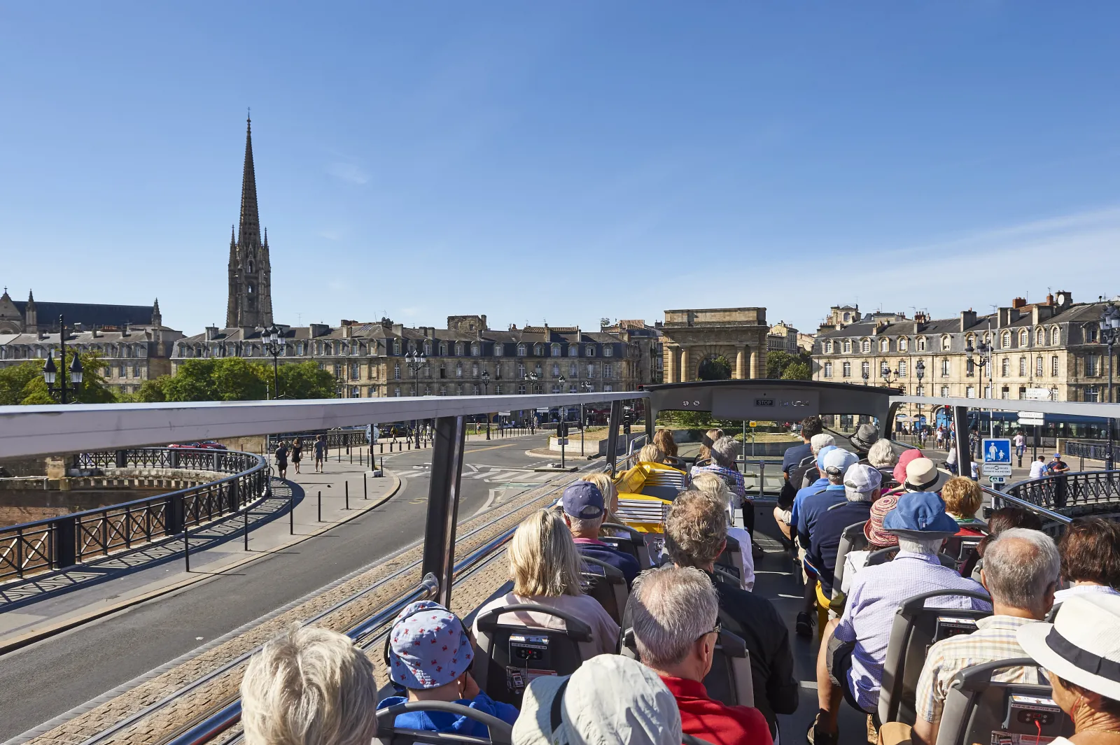 Open-top tour bus passengers enjoying a sightseeing ride through Bordeaux streets, with Haussmann-style buildings and a church spire visible in the background.