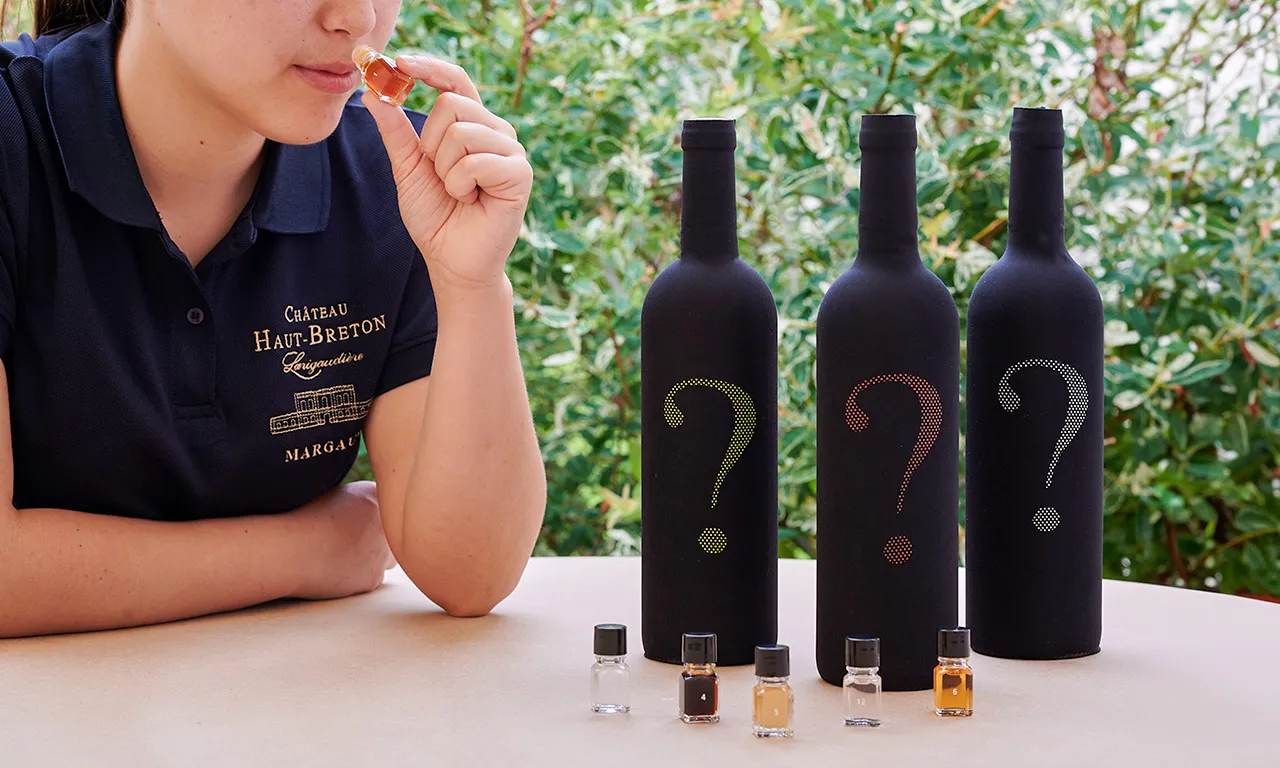 A woman in a Château Haut-Breton polo shirt tastes red wine in front of three mysterious bottles marked with question marks, with tasting glasses on the table.