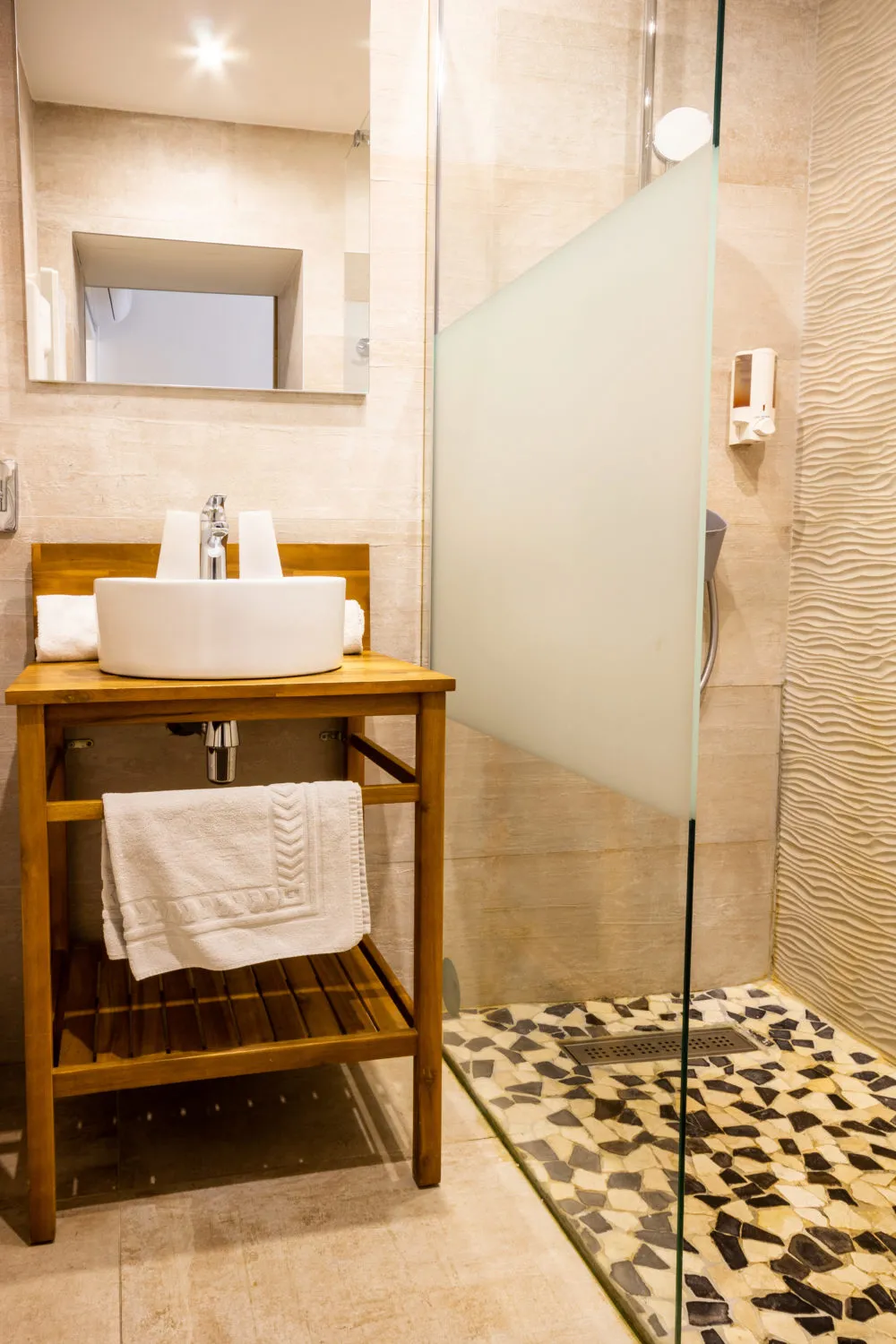 Modern bathroom featuring a wooden vanity with round white basin, illuminated mirror, walk-in shower with glass partitions and geometric black and white tile flooring