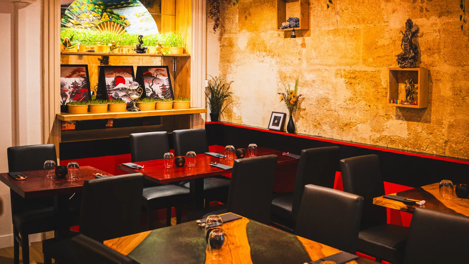 Modern restaurant interior in Bordeaux featuring golden stone walls, black and red furnishings, comfortable seating booths, wooden tables, decorative shelves with plants and Asian artwork, warm and sophisticated atmosphere.