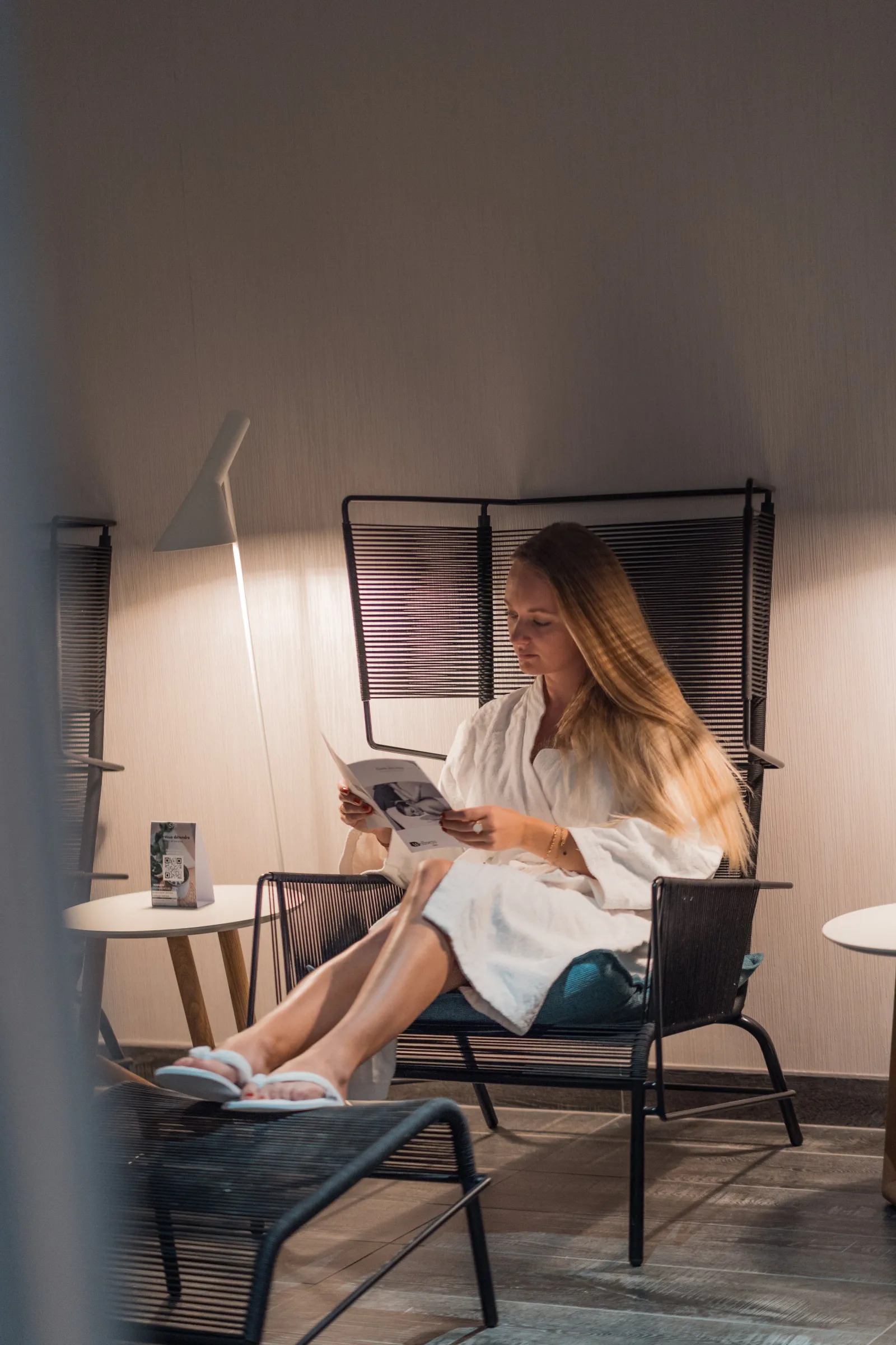 A long-haired woman sitting in a black metal-framed armchair wearing a white shirt, reading a magazine in a modern room at Radisson Blu Hotel Bordeaux. A sleek floor lamp illuminates the space, with a small side table nearby.