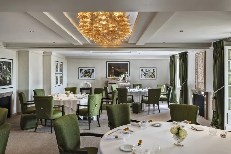 Elegant dining room with round tables set for service, green velvet chairs, golden chandelier, white columns and green curtains at Lalique restaurant, Château Lafaurie Peyraguey in Bordeaux.