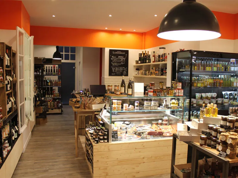 Interior of a gourmet grocery store in Bordeaux featuring wooden counters, refrigerated display cases, shelves of regional products, orange and white walls, and a large black hanging light