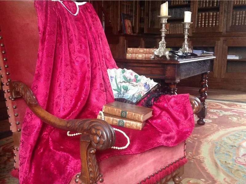 Interior of Château de Vayres near Bordeaux: carved wooden armchair upholstered in bright pink velvet, with antique books and pearl necklace, in a dark wood-paneled library
