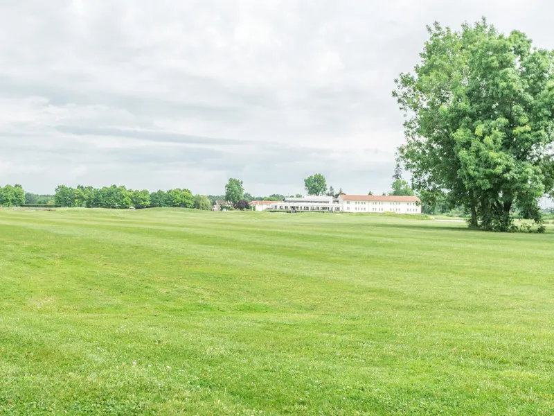Panoramic view of Golf de Margaux near Bordeaux featuring a vast green fairway, majestic trees, and a stone building in the distance under a cloudy sky.