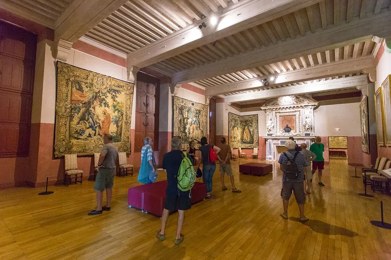 Interior of Château ducal de Cadillac with visitors viewing antique tapestries on walls, light wooden coffered ceiling, wooden parquet floor, period furniture and Renaissance decor.